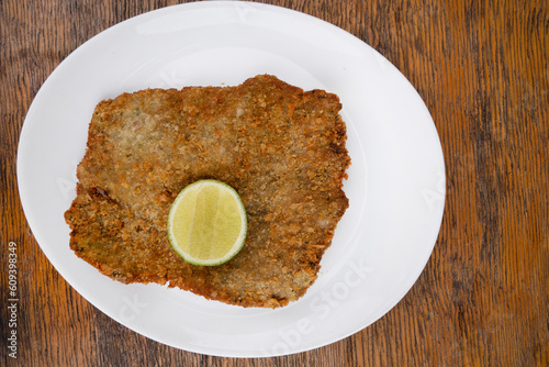 Argentine dish. Top view of milanesa, breaded fried steak, with a slice of lemon, in a white dish with a wooden background. 