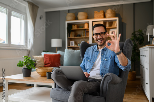 Foto Portrait of cheerful male freelancer with laptop sitting comfortably on armchair and showing OK sign