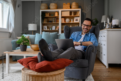 Portrait of cheerful young businessman laughing and doing research over laptop while relaxing legs on footstool. Male entrepreneur working over computer while sitting comfortably on armchair