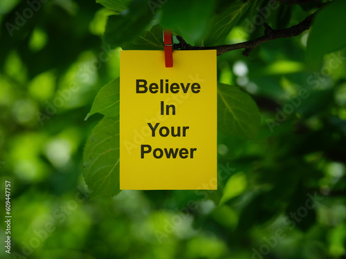 A yellow paper note with the words Believe in Your Power on it attached to a tree branch with a clothes pin