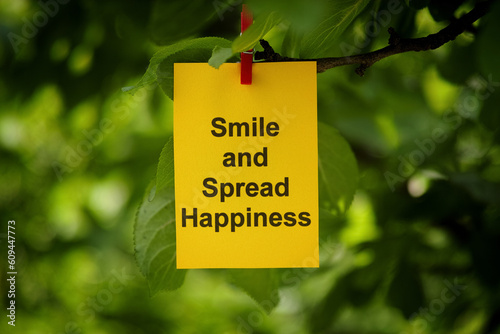 A yellow paper note with the words Smile and Spread Happiness on it attached to a tree branch with a clothes pin © Stepan Popov