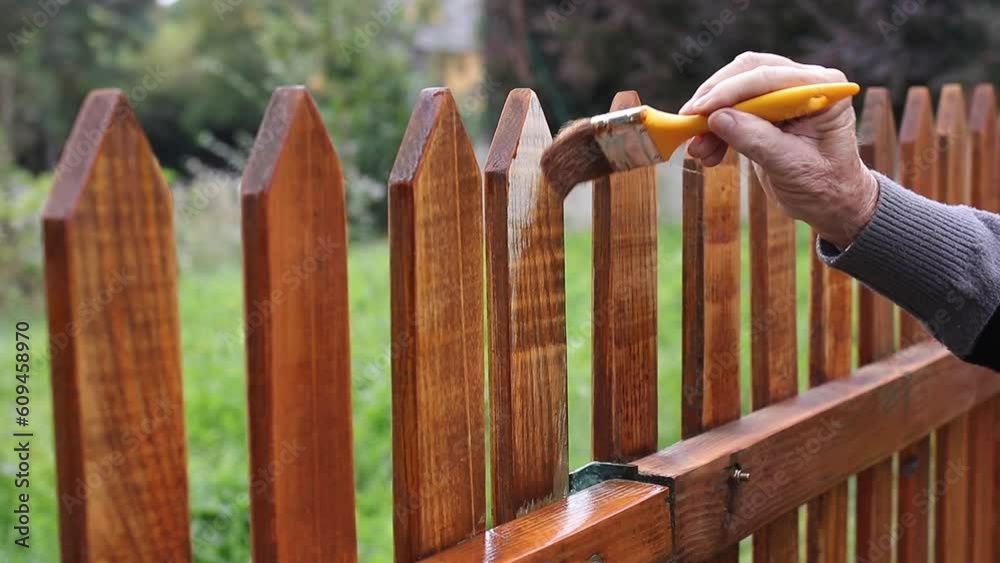 Painting protective varnish on wooden picket fence at backyard. Motion of hand with paintbrush. Handheld shot