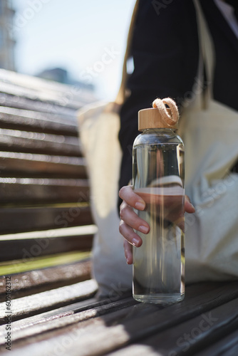 Young woman sitting on a bench in the morning and holding a glass water bottle in hand. Unrecognizable female person chilling outdoor in a sunny day with an eco bottle of fresh water