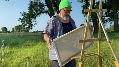 Cool trendy mature artist man preparing to create artwork looks around for best landscape view to paint picture outdoors in morning sunshine. Painter starts to work installs canvas on easel at field photo