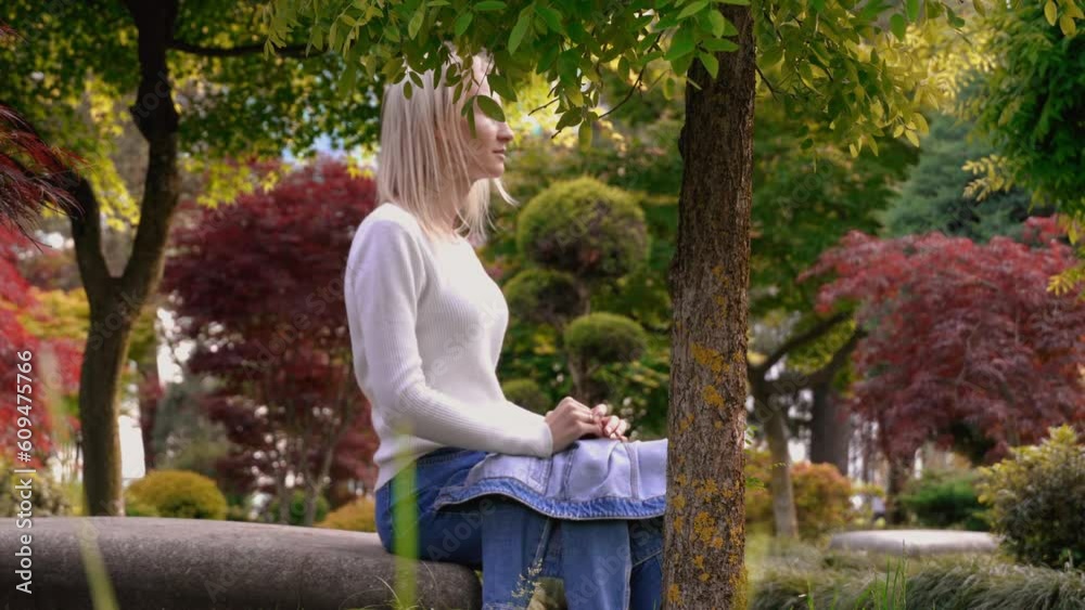 Charming and pretty young girl using phone and relaxing, sitting on bench in beautiful green park on sunny warm day. She smiles and looks into telphone. blonde.