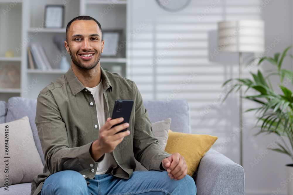 Portrait of a Latin American man sitting at home on the sofa and using the phone. He looks at the camera with a smile Portrait of a Latin American man sitting at home on the sofa and using the phone. He looks at the camera with a smile