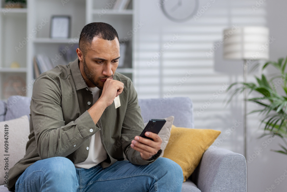 A young African American man received bad news, a message on his cell phone. He sits upset at home on the sofa, looks at the screen in shock