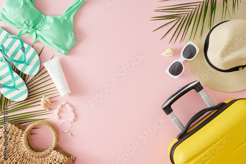 Sandy summer getaway concept. Top view flat lay of sunscreen bottle, yellow suitcase, beach accessories, palm leaves, seashells on pastel pink background with blank space for branding or promotion