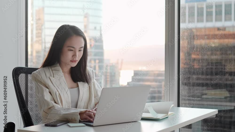 Young busy happy Asian business woman manager using laptop and mobile cell phone devices in office. Professional female executive holding smartphone, working on computer sitting at desk. Young busy happy Asian business woman manager using laptop and mobile cell phone devices in office. Professional female executive holding smartphone, working on computer sitting at desk.