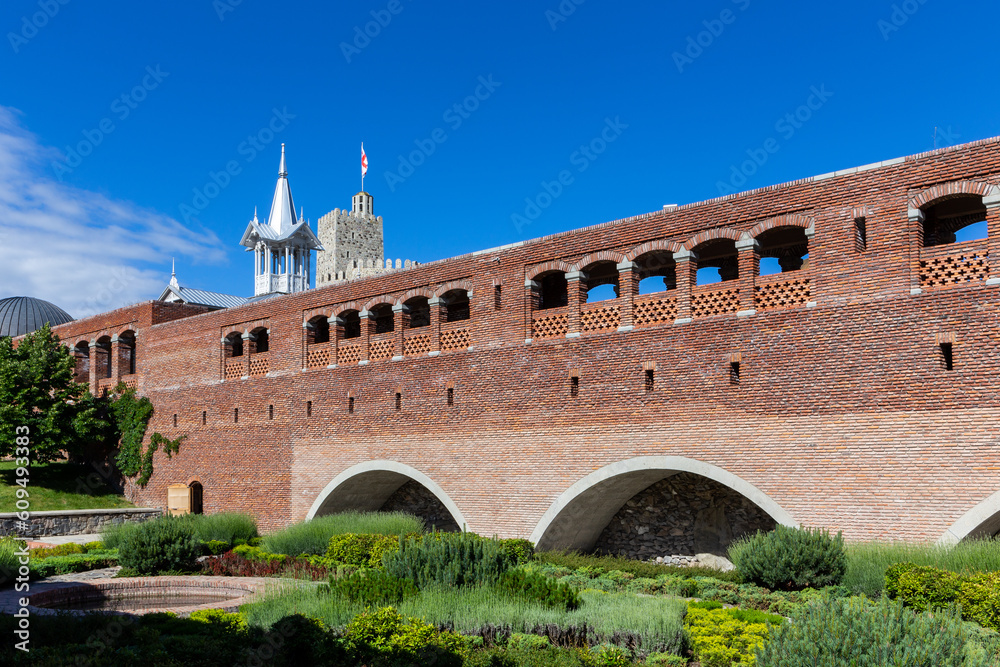 Fototapeta premium Red stone and brick bridge leading to Akhaltsikhe (Rabati) Castle, with cloisters and old moat overgrown with plants, flowers and grass, castle watchtower in the background.