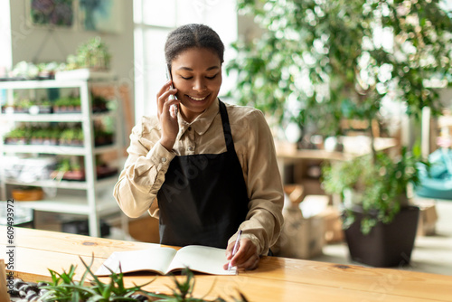 Black female gardener noting client order during mobile phone conversation, workin in greenhouse. Successful botanist and store owner concept