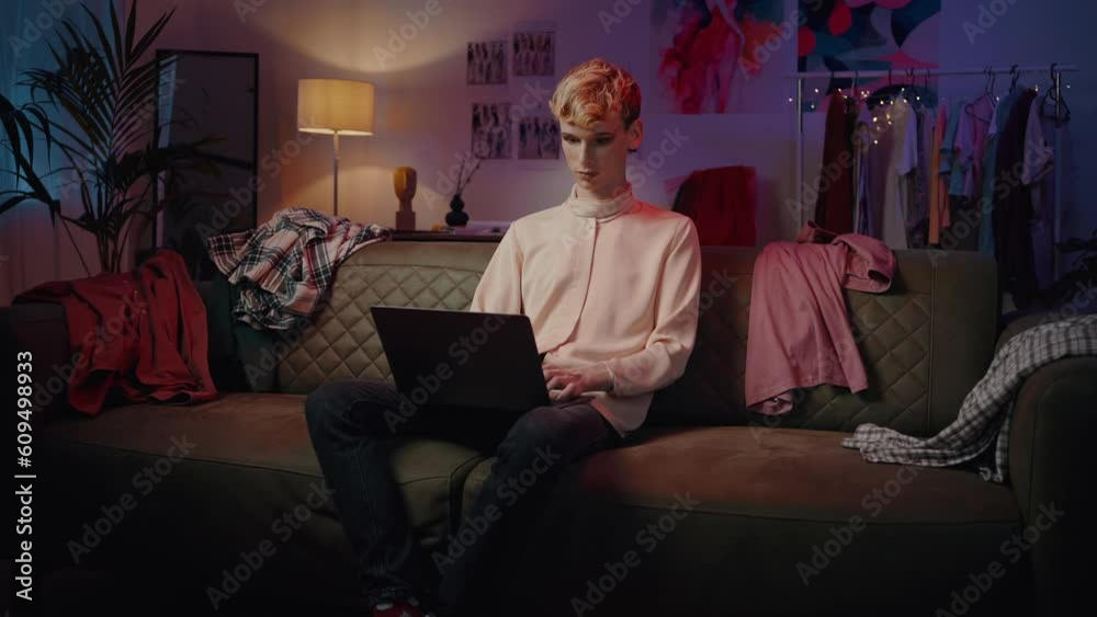 Young non-binary guy sitting on the couch, working remotely on laptop at home with neon lights
