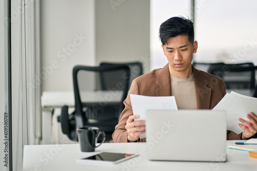 Serious busy young Asian professional business man executive ceo manager sitting at desk in office working checking corporate financial accounting documents feeling worried about taxes or bills.