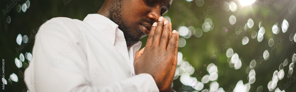 Peaceful African Man Praying In White Shirt Holding Clasped Hands In Prayer Gesture Standing With Eyes Closed Posing In Green Nature Background. Portrait Of Black Man Sending Prayers To God Peaceful African Man Praying In White Shirt Holding Clasped Hands In Prayer Gesture Standing With Eyes Closed Posing In Green Nature Background. Portrait Of Black Man Sending Prayers To God