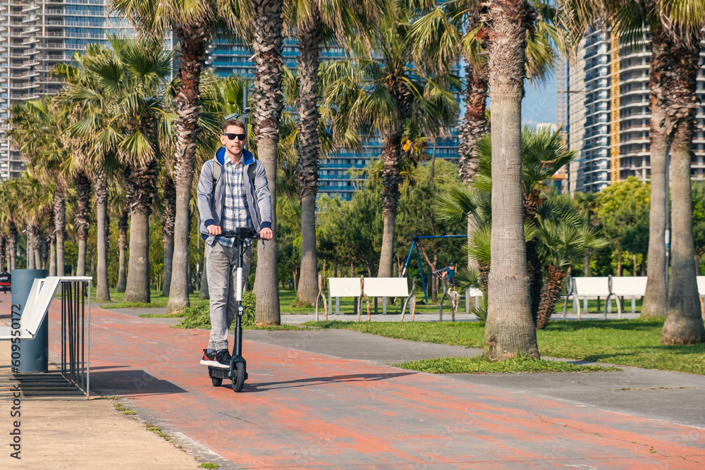 Young man riding electric scooter eco friendly transport along the promenade in the city with palm trees. Male driving e-scooter sustainable and outdoor in summer day. Sustainable lifestyle Young man riding electric scooter eco friendly transport along the promenade in the city with palm trees. Male driving e-scooter sustainable and outdoor in summer day. Sustainable lifestyle