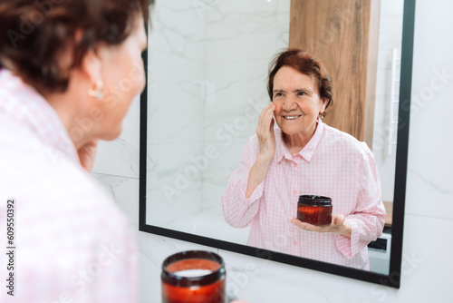Senior mature happy woman, older lady looking at mirror applying whitening antiaging anti wrinkle perfect skin care cream to eye contour