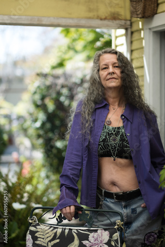 An LGBTQ senior poses in their backyard. They are showing their bare midriff and face piercings, expressing fun and vibrant queer expression. Their long silver hair suggests their maturity and age.