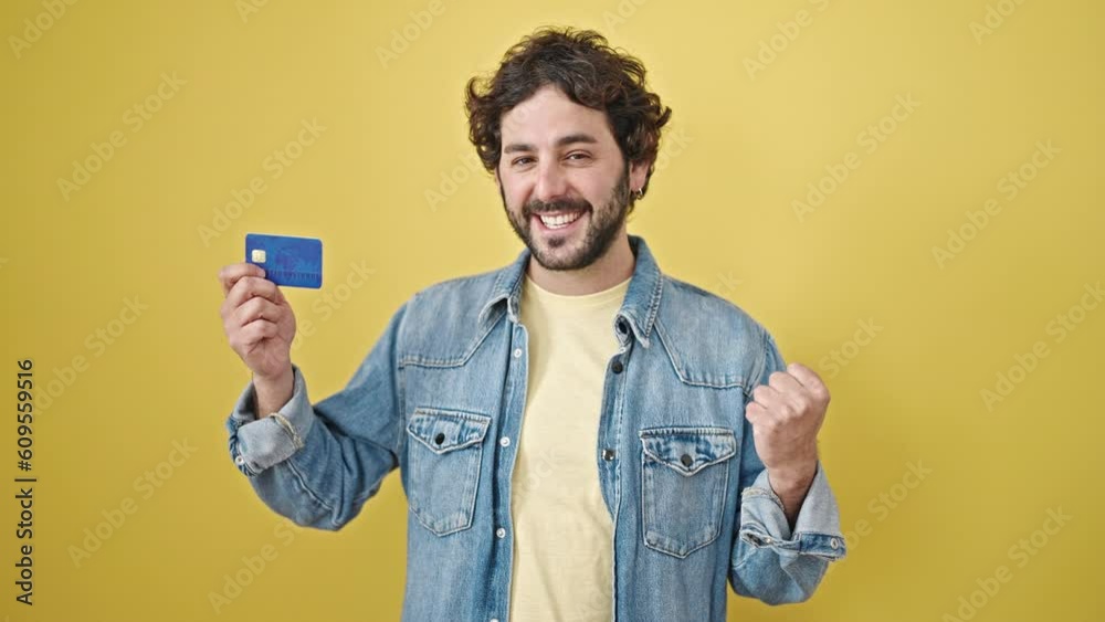 Young hispanic man holding credit card with winner expression over isolated yellow background