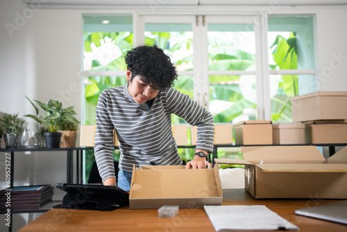 Front view of a young short curly hair Asian female small business owner, standing at desk talking on the phone holding between her ear and shoulder while checking the product in the box with a tablet