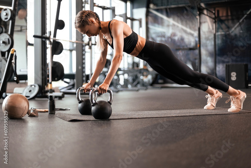 Portrait of a muscular woman on a plank position with kettlebell at gym.