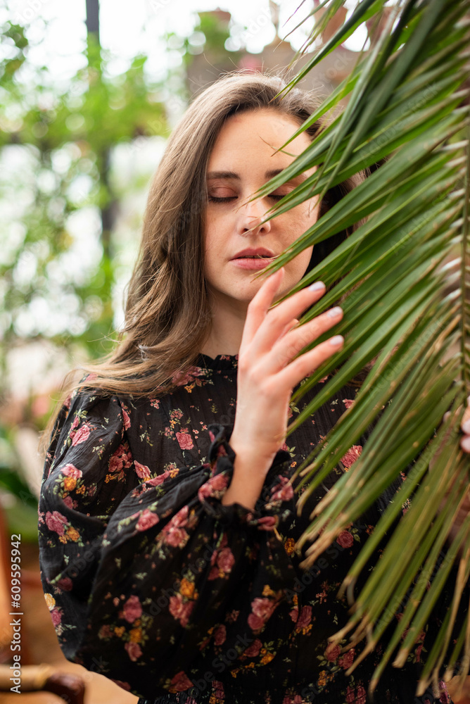 Young beautiful woman in black dress with long brown hair holding big palm leaf in greenhouse Young beautiful woman in black dress with long brown hair holding big palm leaf in greenhouse
