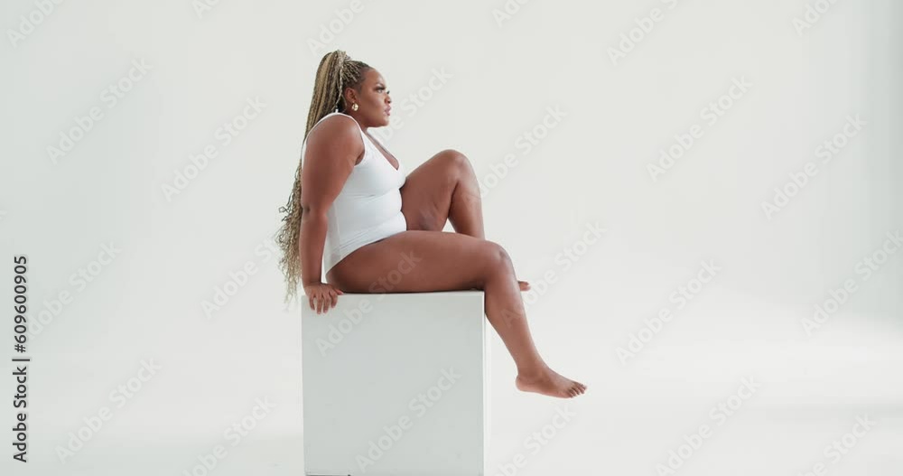 Body positive plus size clumsy female sitting on white cube. Woman in bodysuit is posing in the studio. Afro girl with dreads has rest, break, looking at camera, smiling free spare time