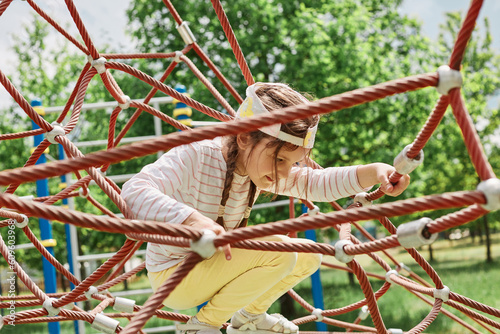 Funny little girl with braids in cap climbing, playing with ropes. summer activity in park in camp having fun alone healthy holiday activity for children.