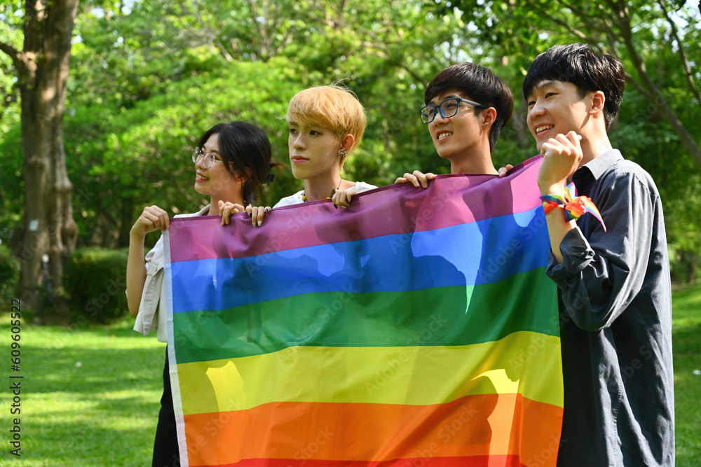 Portrait with group of young LGBT teenagers holding pride flag together with nature park on the background.
