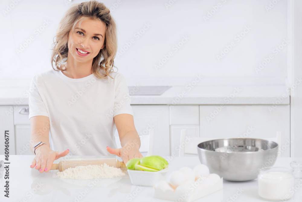 A beautiful young woman is preparing dinner at the kitchen table, looking at the camera and smiling. Caucasian blonde is engaged in cooking. Portrait of a happy housewife in a bright home kitchen.