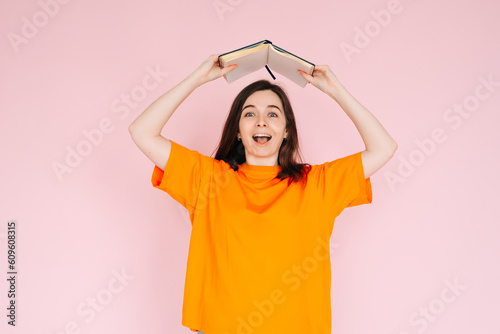 Joyful Book Enthusiasm: Cheerful Woman Holding Book Overhead with Exuberance - Delightful Mood and Positivity Concept, Isolated on Pink Background © Yauhen
