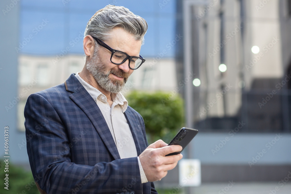 A successful gray-haired businessman uses phone outside an office building, a senior boss in a business suit walks down the street, a man reads messages and browses online pages, uses an application. A successful gray-haired businessman uses phone outside an office building, a senior boss in a business suit walks down the street, a man reads messages and browses online pages, uses an application.