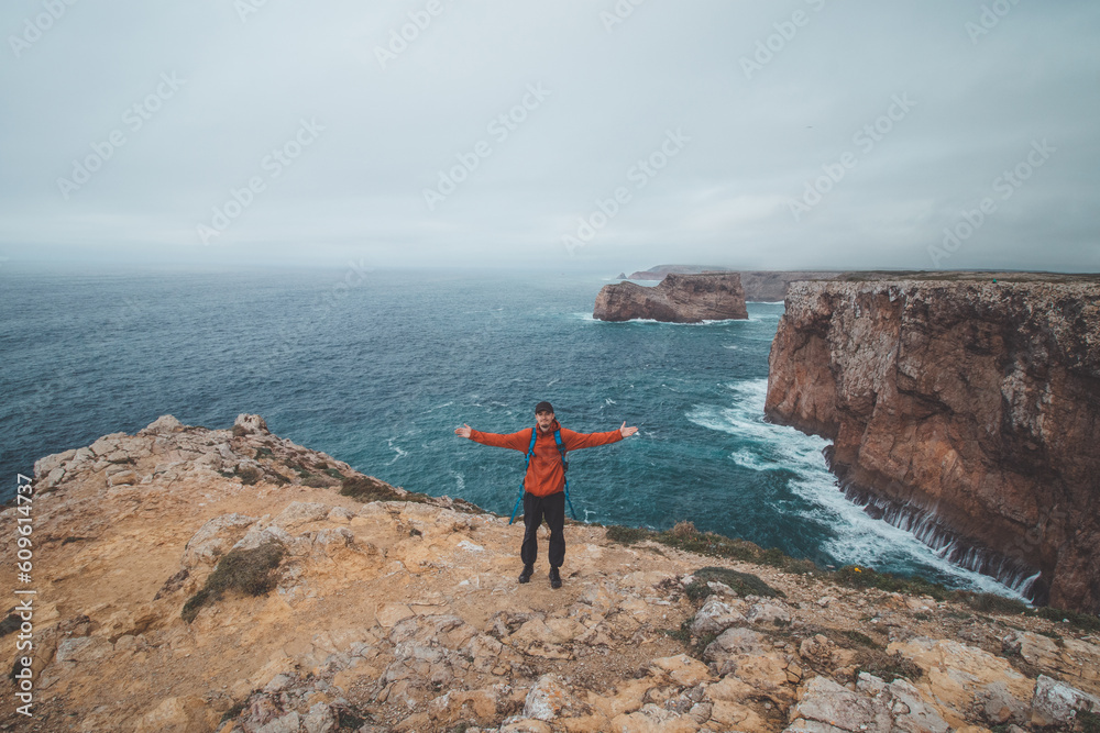 Fototapeta premium Black-haired adventurer stands at the end of Cape Cabo de Sao Vicente in the southwest of Portugal in the Algarve region. Man is enjoying his freedom. Wandering of Fisherman Trail