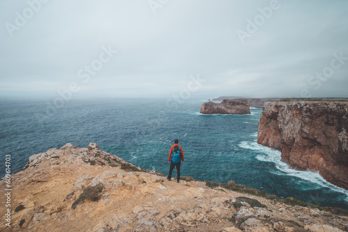 Black-haired adventurer stands at the end of Cape Cabo de Sao Vicente in the southwest of Portugal in the Algarve region. Man is enjoying his freedom. Wandering of Fisherman Trail © Fauren