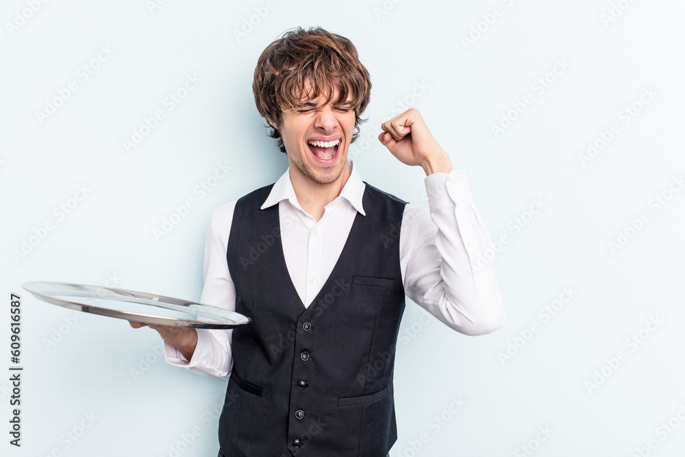 Young caucasian waitress man holding a tray isolated on blue background raising fist after a victory, winner concept. Young caucasian waitress man holding a tray isolated on blue background raising fist after a victory, winner concept.
