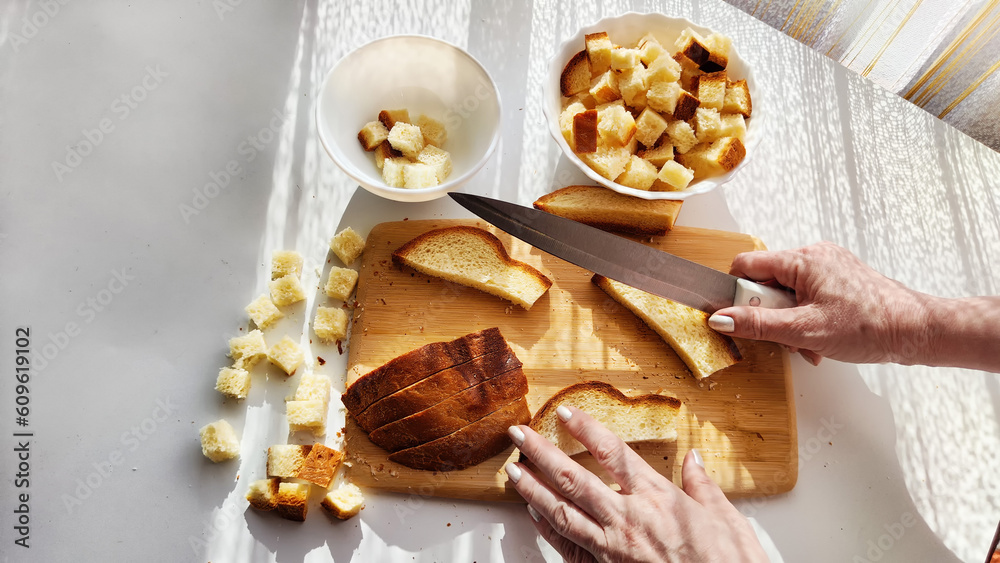 Fototapeta premium Homemade cooking and cutting of slices of diced dry white rye bread on cutting board and white plate. Hand of woman with Crackers for food, healthy eating on table in sunny day, top view