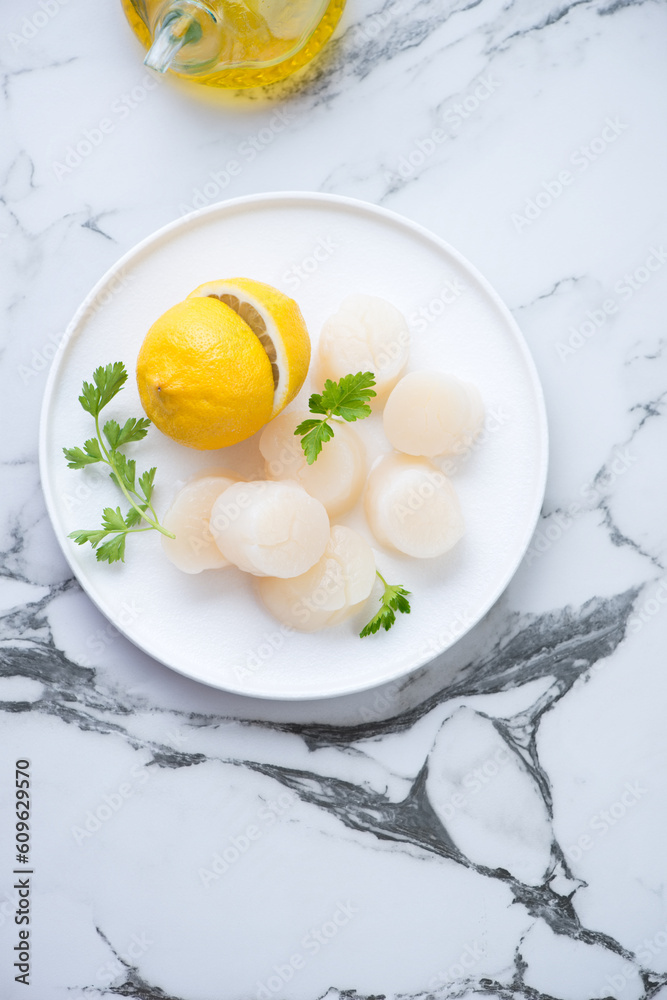 Plate with fresh uncooked sea scallops, lemon and parsley, vertical shot on a white marble background with space, flatlay Plate with fresh uncooked sea scallops, lemon and parsley, vertical shot on a white marble background with space, flatlay