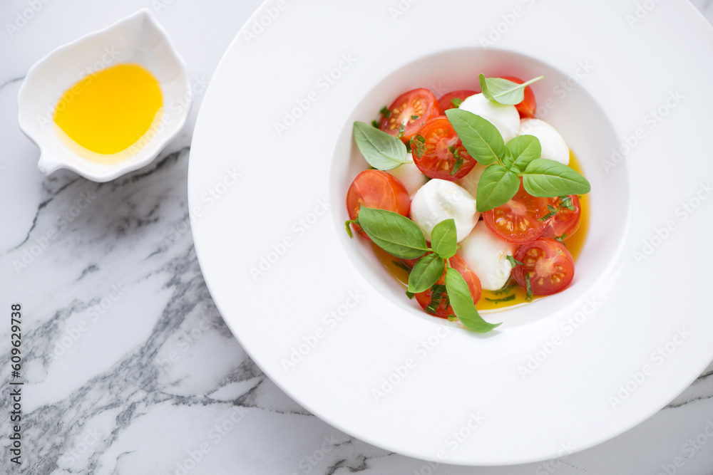 Caprese with mini mozzarella, red cherry tomatoes, olive oil and green basil served in a white plate, middle close-up, high angle view