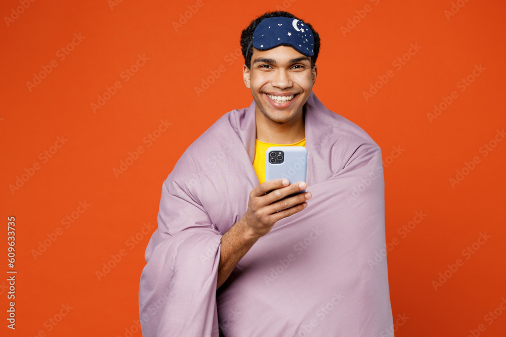 Calm young fun man wear pyjamas jam sleep eye mask wrapped in purple blanket use mobile cell phone rest relax at home isolated on plain orange background studio portrait. Good mood night nap concept. Calm young fun man wear pyjamas jam sleep eye mask wrapped in purple blanket use mobile cell phone rest relax at home isolated on plain orange background studio portrait. Good mood night nap concept.
