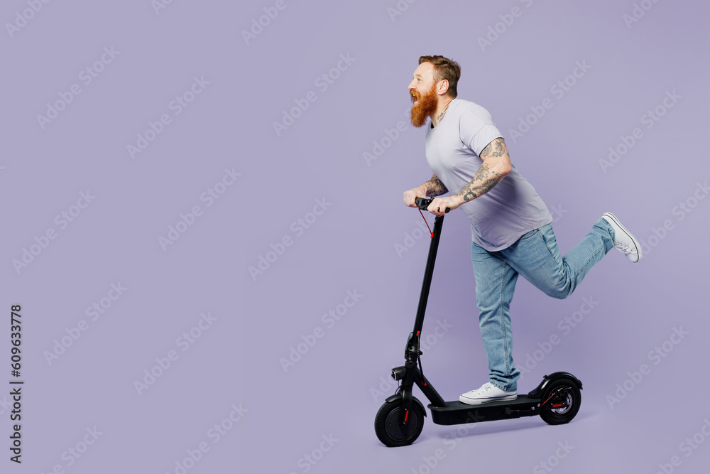 Full body happy fun young redhead bearded man he wears violet t-shirt casual clothes rising e-scooter raise up leg isolated on plain pastel light purple background studio portrait. Lifestyle concept. Full body happy fun young redhead bearded man he wears violet t-shirt casual clothes rising e-scooter raise up leg isolated on plain pastel light purple background studio portrait. Lifestyle concept.