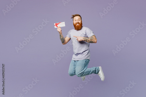 Full body young redhead bearded man wear violet t-shirt casual clothes jump high hold gift certificate coupon voucher card for store isolated on plain pastel light purple background studio portrait.