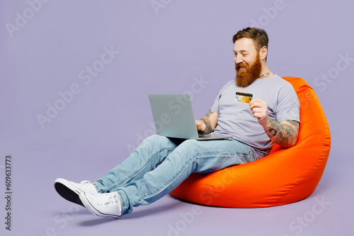 Full body young redhead bearded IT man in violet t-shirt casual clothes sit in bag chair hold use work on laptop pc computer isolated on plain pastel light purple background studio Lifestyle concept