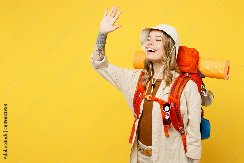 Side view young smiling fun woman carry backpack with stuff mat waving hand isolated on plain yellow background. Tourist leads active lifestyle walk on spare time Hiking trek rest travel trip concept Side view young smiling fun woman carry backpack with stuff mat waving hand isolated on plain yellow background. Tourist leads active lifestyle walk on spare time Hiking trek rest travel trip concept