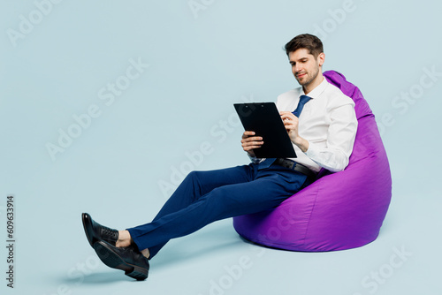 Full body young employee IT business man corporate lawyer wear classic formal shirt tie work in office sit in bag chair clipboard with paper account documents isolated on plain pastel blue background.