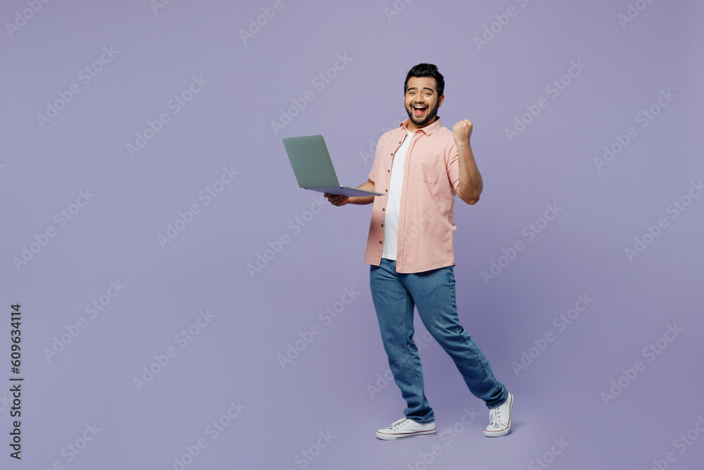 Full body fun young IT Indian man he wear pink shirt white t-shirt casual clothes hold use work on laptop pc computer walk go do winner gesture isolated on plain pastel light purple background studio.