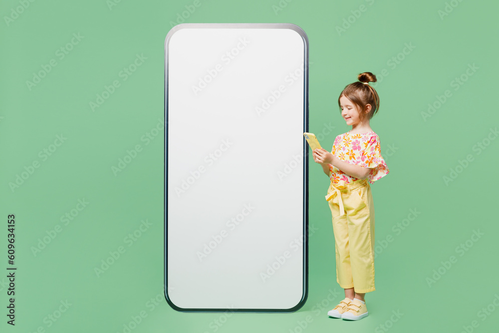Full body side view little child kid girl 6-7 years old wear casual clothes big blank screen area mobile cell phone use smartphone isolated on plain green background. Mother's Day love family concept. Full body side view little child kid girl 6-7 years old wear casual clothes big blank screen area mobile cell phone use smartphone isolated on plain green background. Mother's Day love family concept.