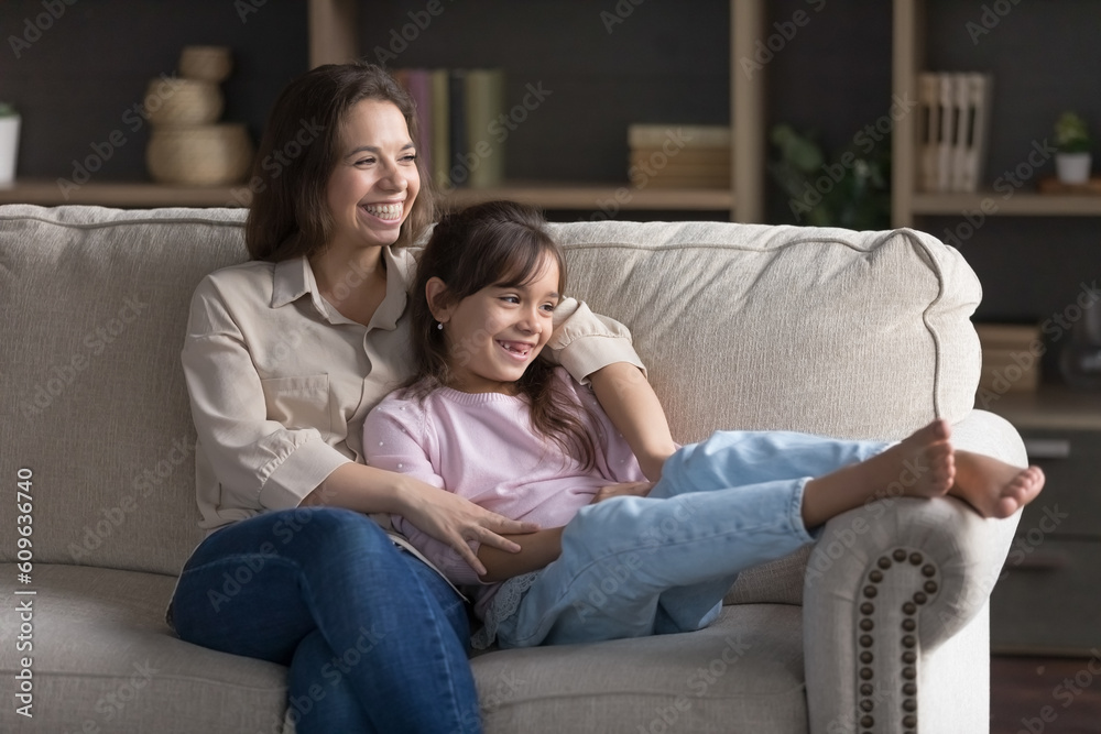 Positive young mom and dreamy sweet daughter kid looking away in happy thoughts, hugging on sofa, resting on home couch together, smiling, laughing, dreaming, thinking on future plans