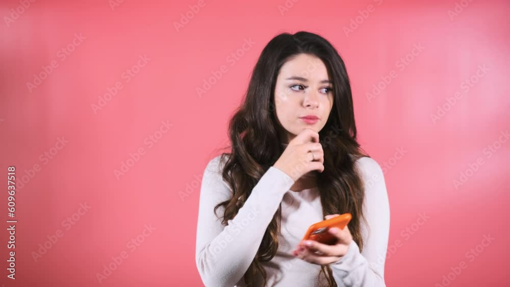 Young woman with amused and smiling expression enjoys scrolling social media on orange smartphone. Long-haired brunette lady smiles looking at feeds Young woman with amused and smiling expression enjoys scrolling social media on orange smartphone. Long-haired brunette lady smiles looking at feeds