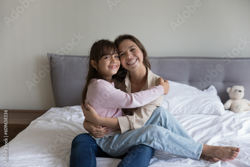 Cheerful pretty mom holding cute preschool kid girl in arms, hugging child, sitting on bed, looking at camera, smiling, enjoying home comfort, motherhood. Mother and little daughter portrait