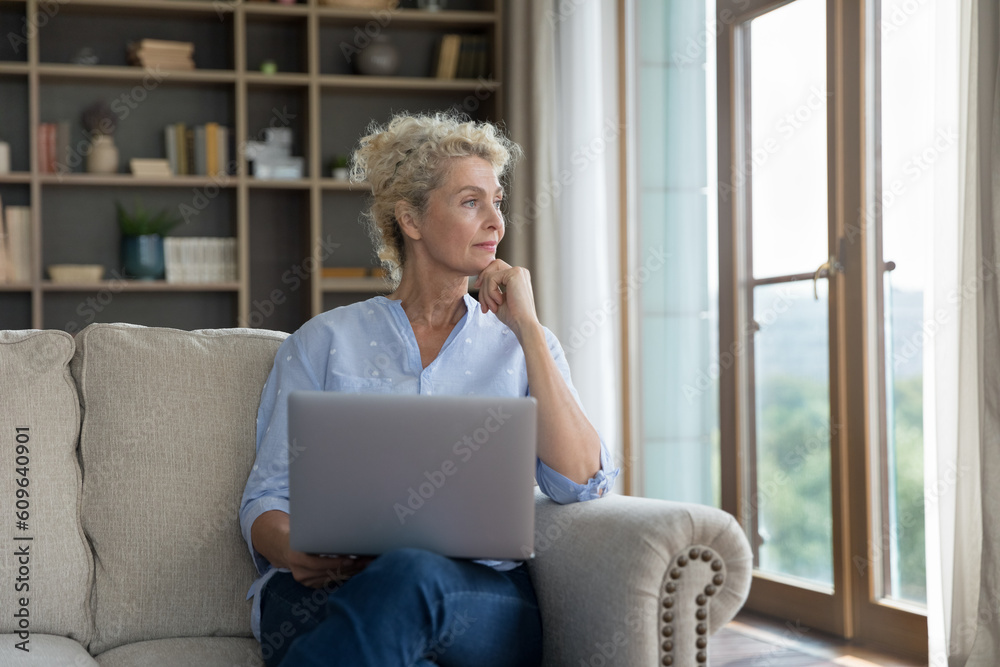Positive pensive senior woman holding laptop on lap, sitting on sofa, looking at window away, toughing chin, thinking, dreaming, planning, using wireless Internet technology for communication Positive pensive senior woman holding laptop on lap, sitting on sofa, looking at window away, toughing chin, thinking, dreaming, planning, using wireless Internet technology for communication
