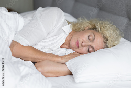 Calm serene sleepy blonde senior woman lying on side, resting on comfortable bed mattress, enjoying relaxation, recreation in morning, sleeping in white linen with head on pillow